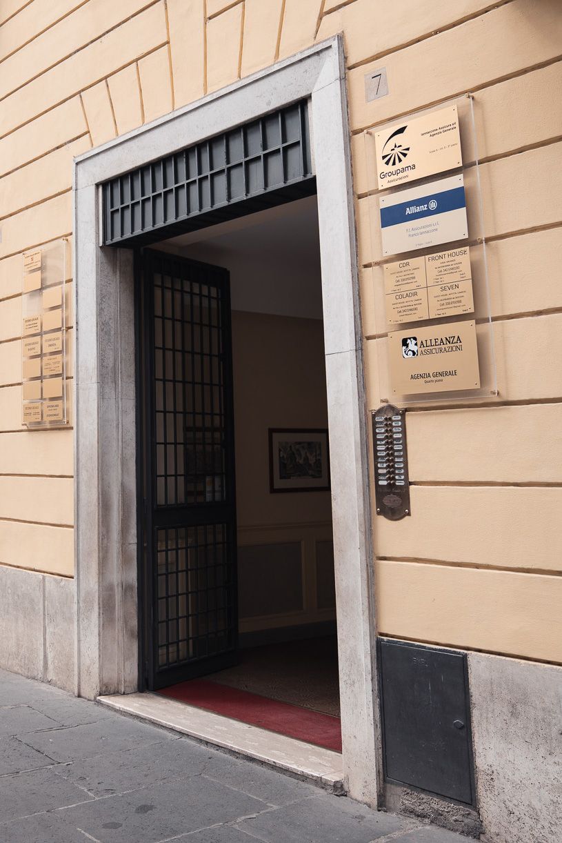 Porta aperta con cancello di sicurezza in metallo; tre cartelli sul muro; edificio color crema.