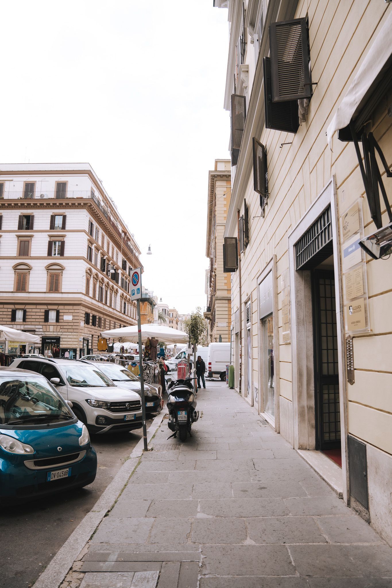 Scena di strada con auto parcheggiate, pedoni ed edifici a Roma. Uno scooter percorre il marciapiede.
