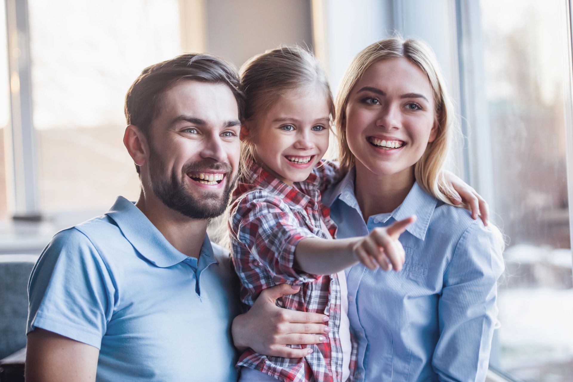Famiglia sorridente, la bambina indica, i genitori la abbracciano vicino alla finestra.