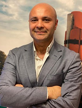 Uomo sorridente con indosso una giacca grigia e braccia incrociate davanti a un espositore di attrezzature di sicurezza marittima.