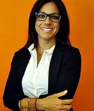 Donna con gli occhiali, blazer nero, camicia bianca, sorridente, braccia incrociate, davanti a uno sfondo arancione.
