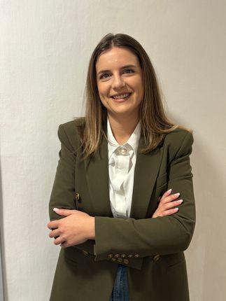 Donna con giacca verde oliva e camicia bianca, braccia incrociate, sorridente.