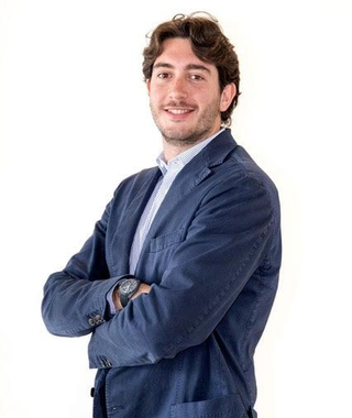 Uomo con i capelli castani, braccia incrociate, indossa un blazer blu navy e una camicia azzurra, sorridente.