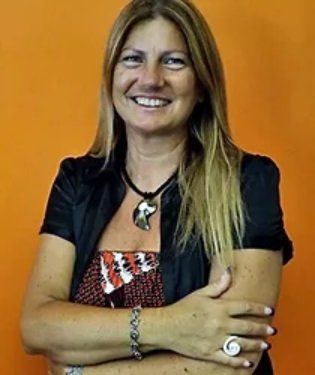 Donna con i capelli biondi, sorridente, braccia incrociate, che indossa una giacca nera e un top fantasia su uno sfondo arancione.