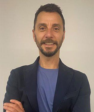 Un uomo con barba e capelli castani corti sorride, con le braccia incrociate, e indossa una camicia blu e un blazer scuro.
