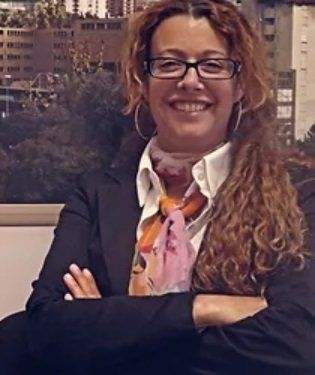 Una donna con occhiali e blazer sorride con le braccia incrociate e indossa una sciarpa fantasia.