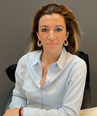 Donna con una camicia azzurra, che guarda la telecamera e indossa orecchini a cerchio, in un ambiente d'ufficio.