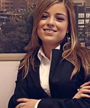 Donna in blazer sorride, braccia incrociate; camicia bianca; sfondo della città.