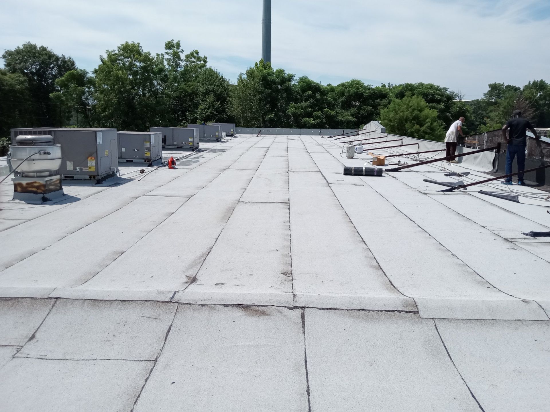 Flat commercial rooftop with white membrane, HVAC units, and two workers inspecting near the edge.