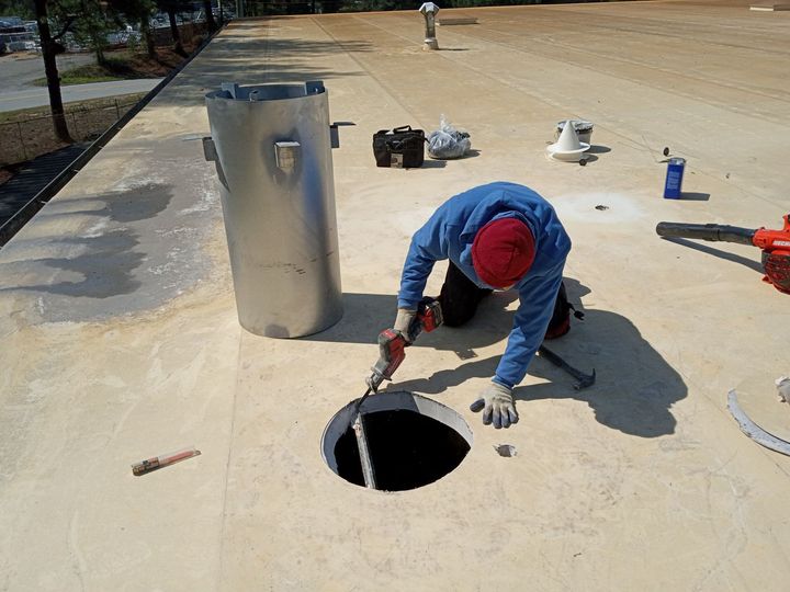 Worker kneeling on a rooftop beside an open vent, using a tool to inspect or repair it.