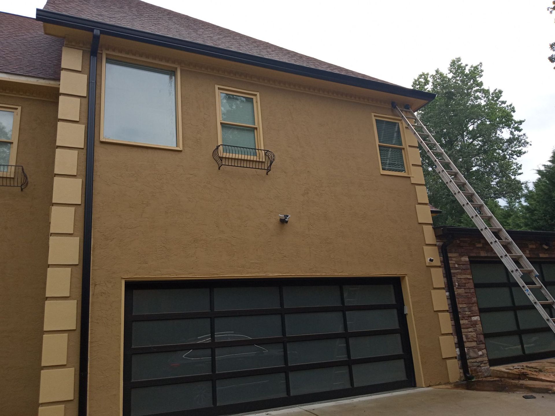 Tan stucco house exterior with garage doors, windows, and a ladder leaning on the right side.