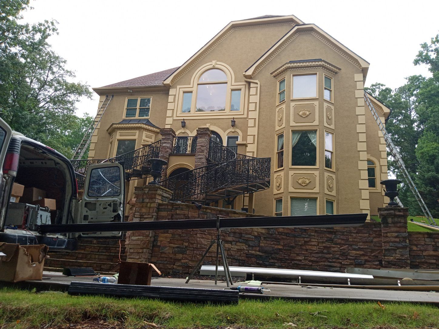 Large beige two-story house with ornate balconies and stone wall, beside a parked truck and excavator