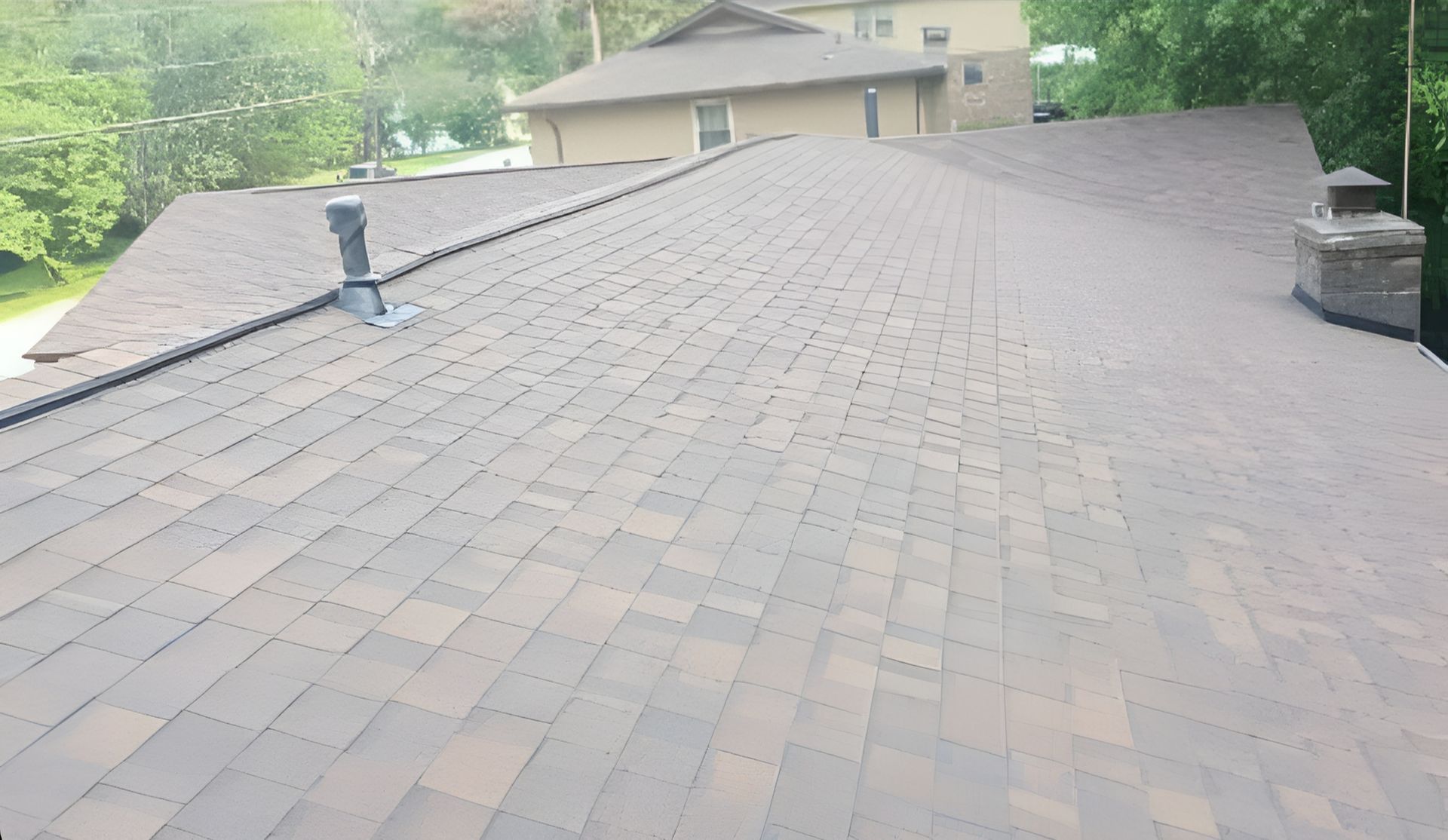 Wide view of a shingled roof with vents and chimneys, seen from above near a house and trees