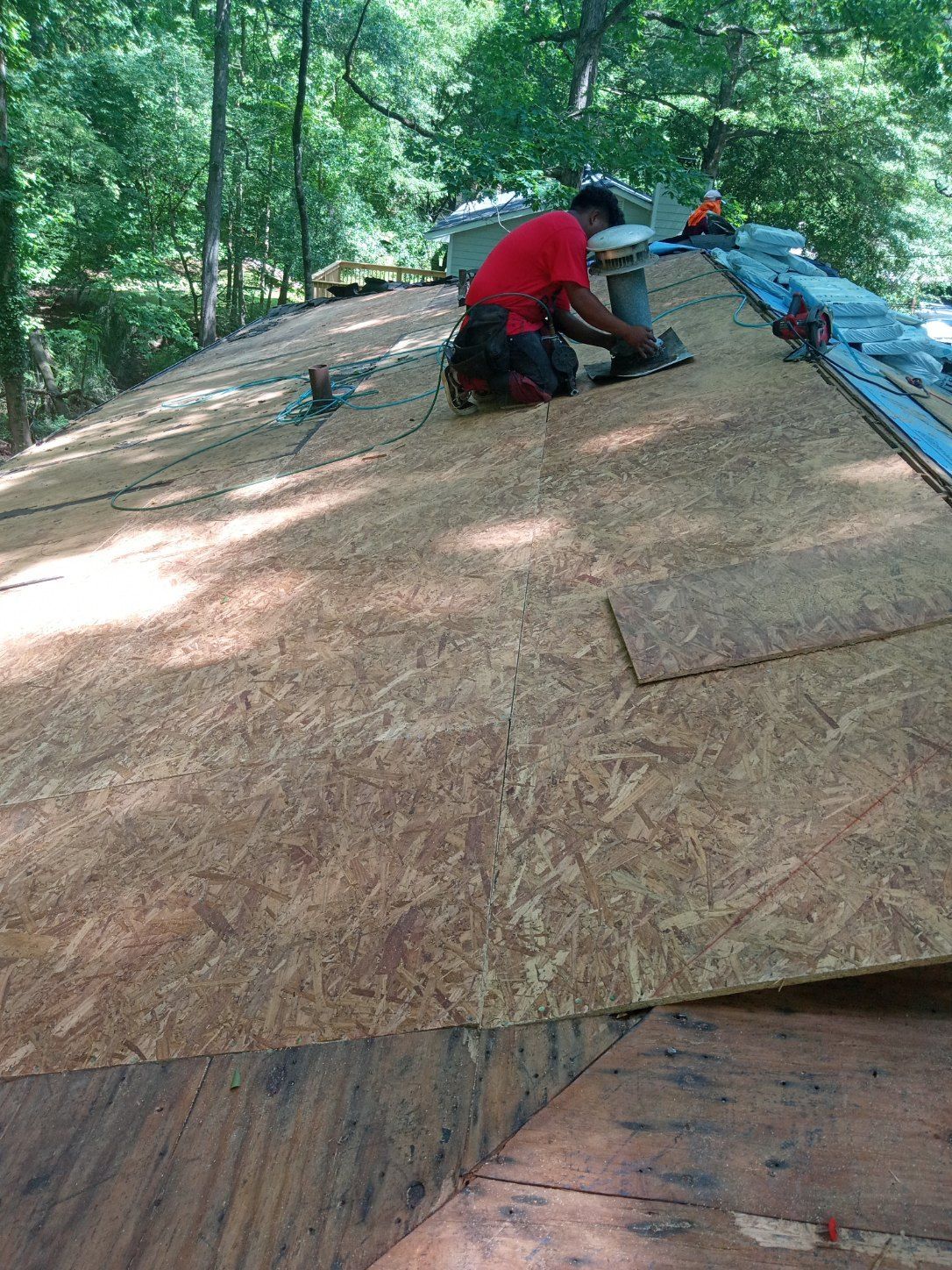 Workers installing plywood roof sheathing on a house in a wooded area, one in a red shirt.