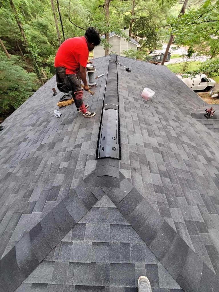 Worker installing shingles on a steep gray roof in a wooded area
