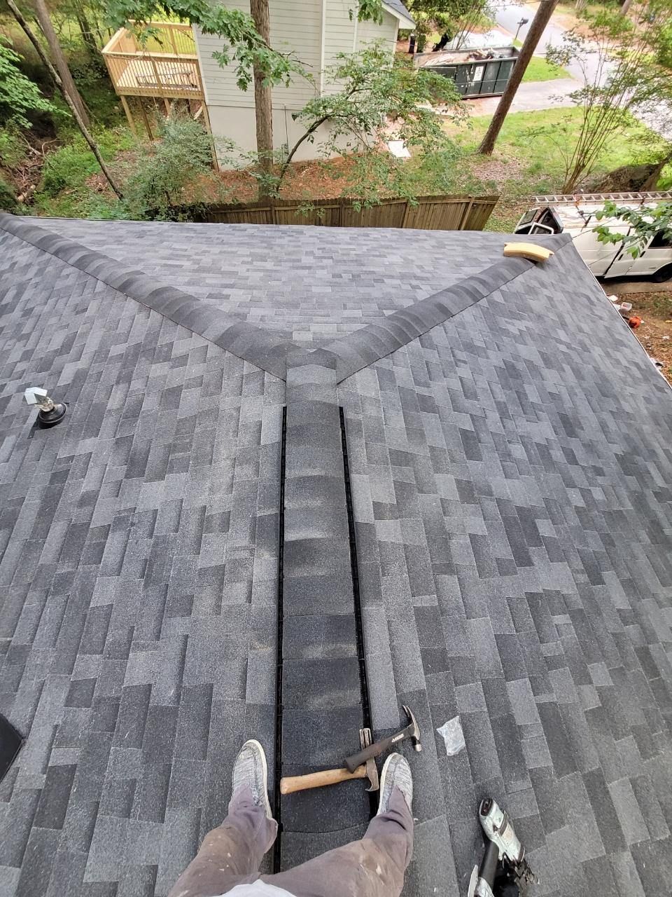 Roofing view from a ladder: gray shingle roof valley with a worker’s boots and tool visible, trees below