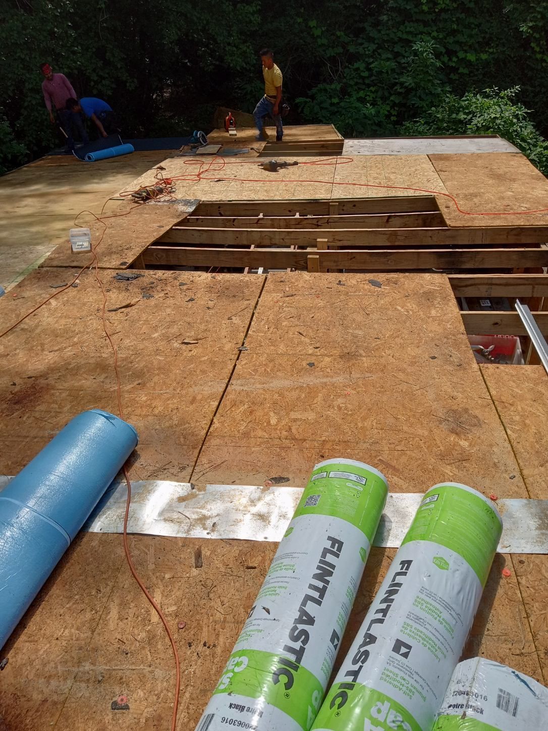 Workers laying plywood sheathing on a rooftop under construction, with insulation rolls in the foreground.