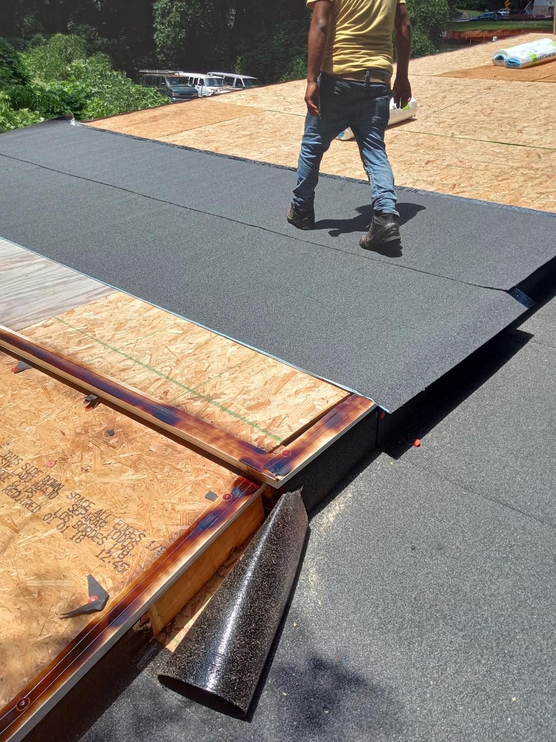 Worker installing black roofing underlayment on a roof, with exposed plywood and flashing visible