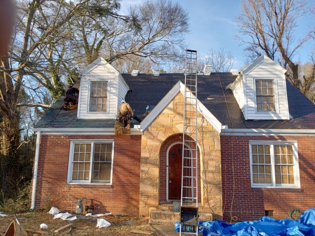 Small brick house with a white roof and dormer windows, surrounded by trees and yard debris.