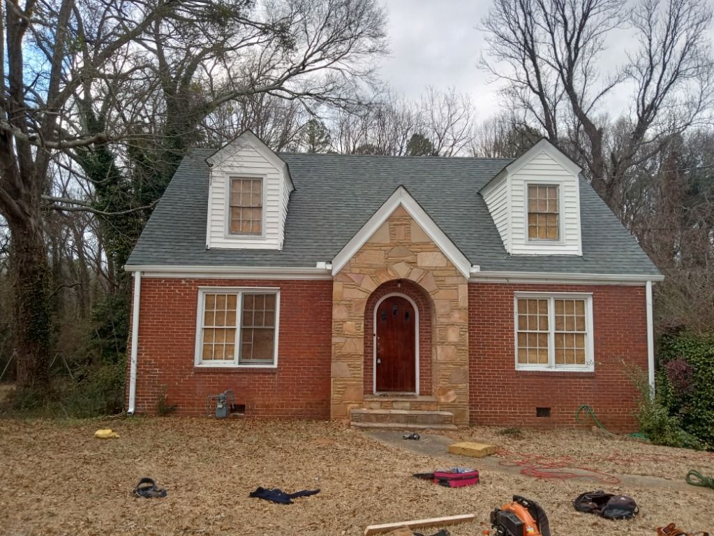 Small brick house with gray roof and dormers, set among bare trees and leaf-covered ground.