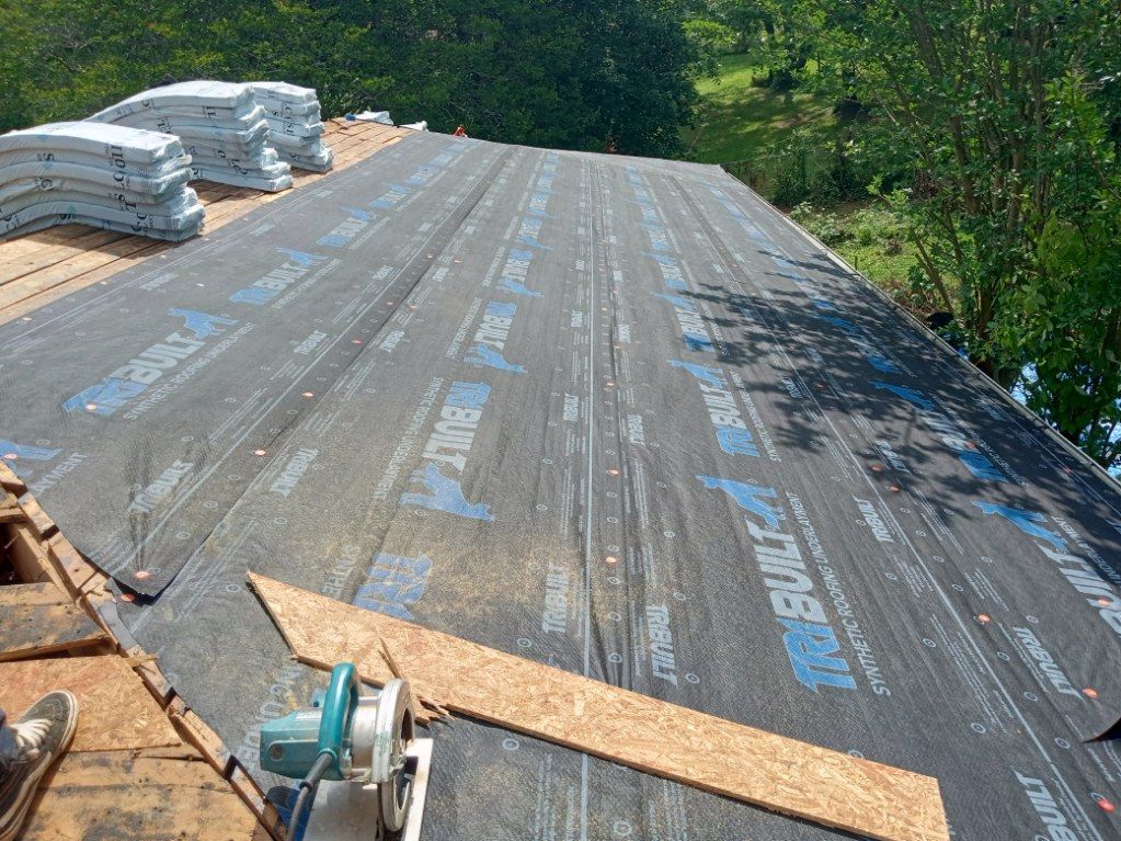 Roof under construction with black underlayment, stacks of shingles, and trees in the background