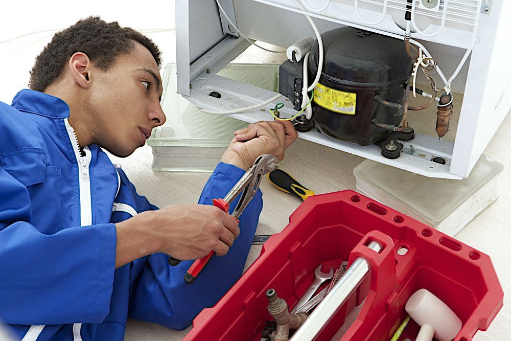 Person in blue overalls fixing a refrigerator compressor with tools from a red toolbox.