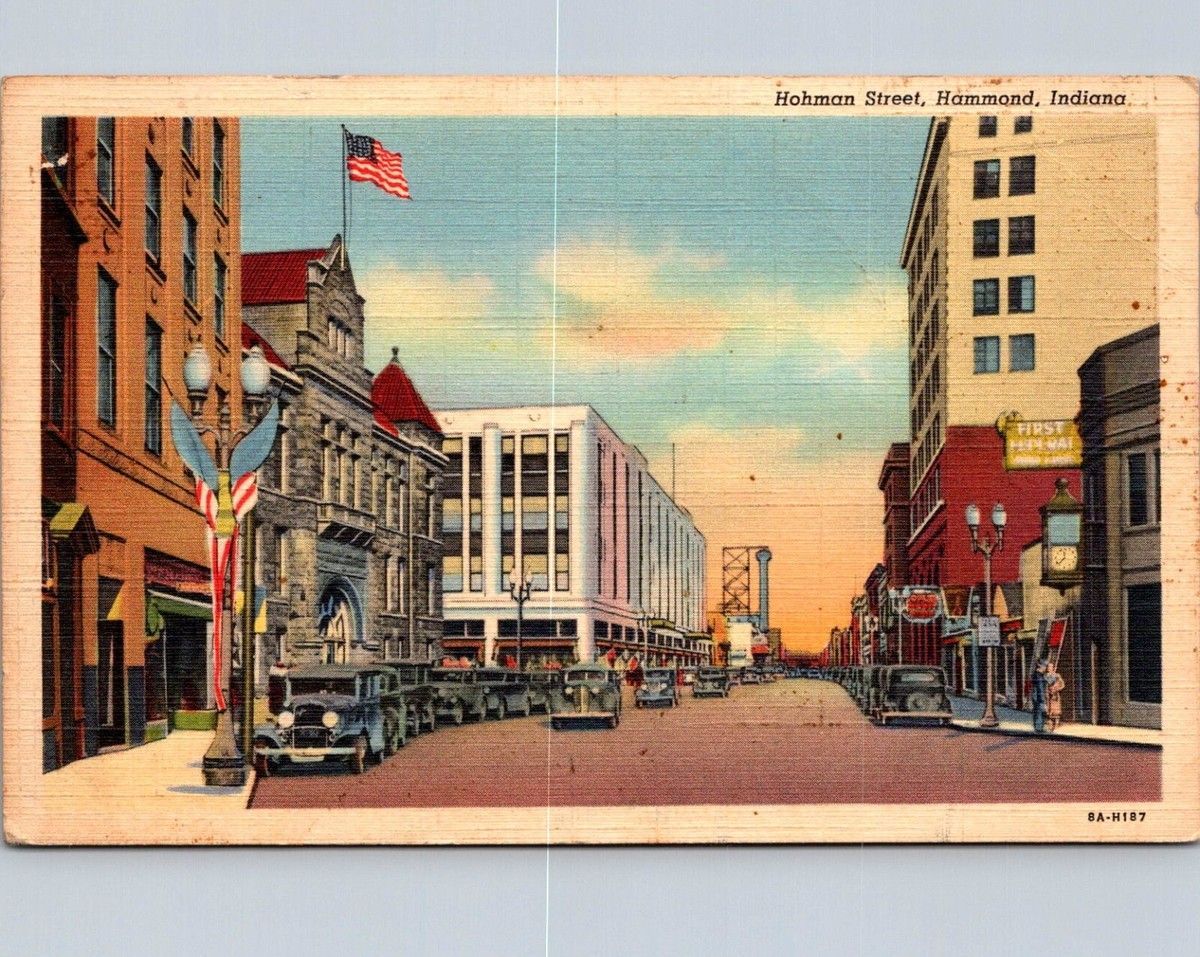 Vintage postcard of a downtown street with cars, brick buildings, and an American flag at sunset.