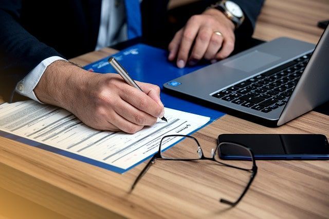 Un hombre firma un documento frente a una computadora portátil.