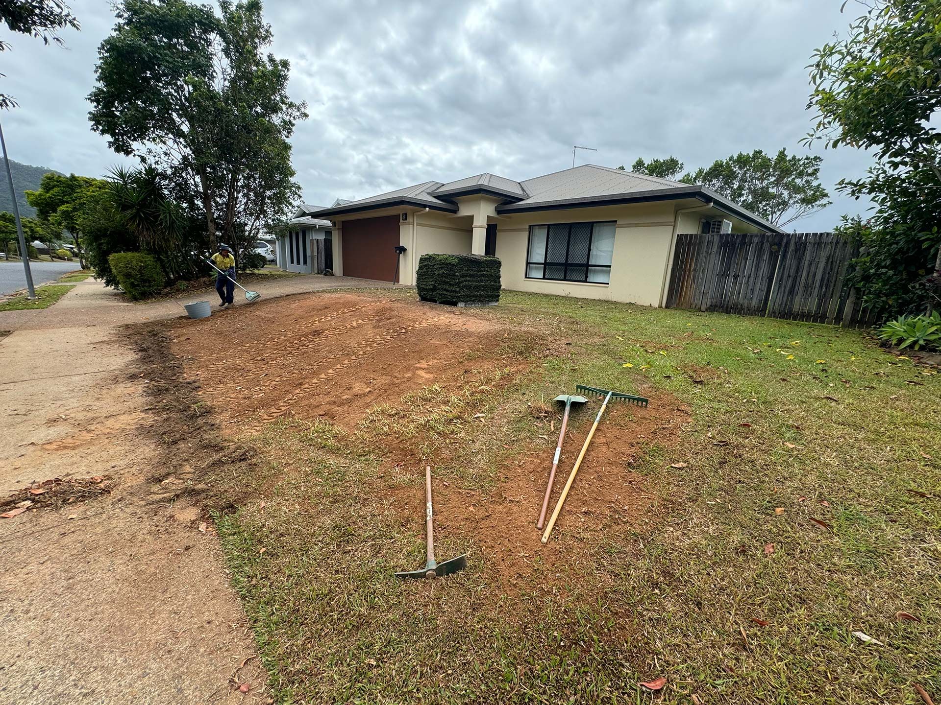 Single-story beige house with a front yard, dirt driveway, and tools on the grass under cloudy skies