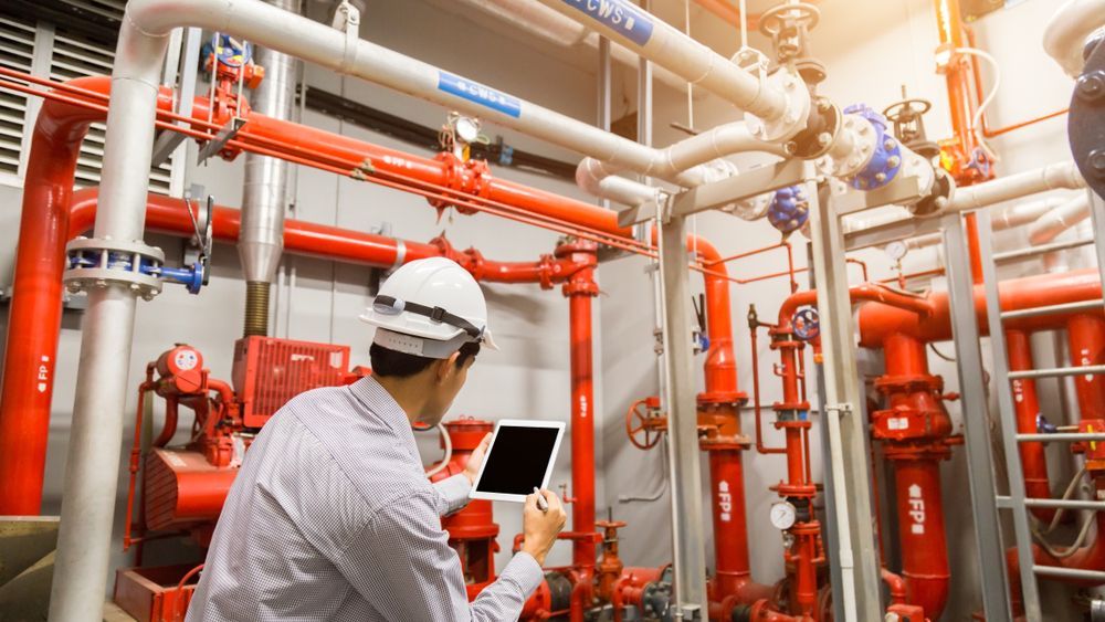 Technician inspecting industrial pipes and valves in a mechanical room with a laptop