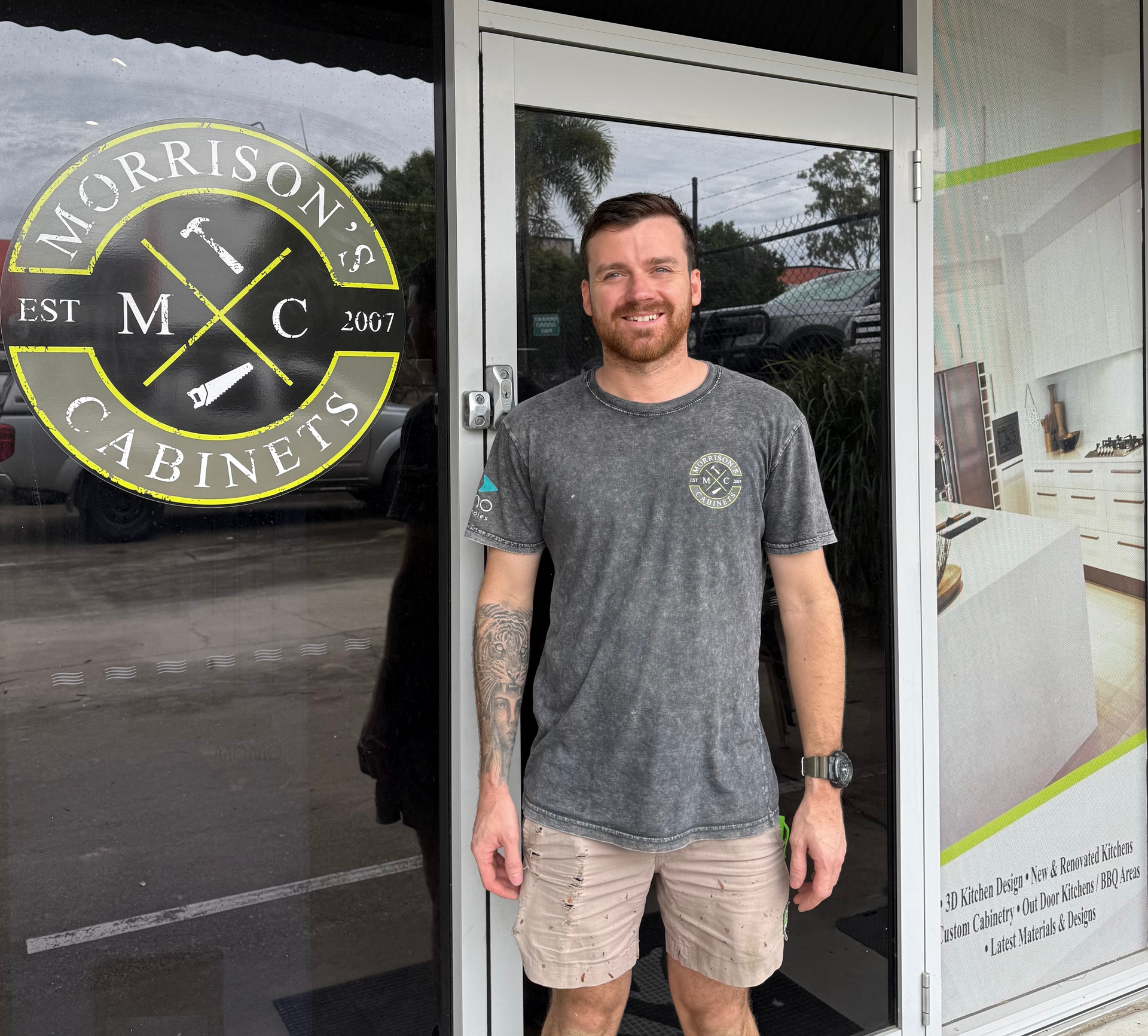 Man standing in a doorway beside a Morrison Cabinet sign, wearing a gray shirt and beige shorts.