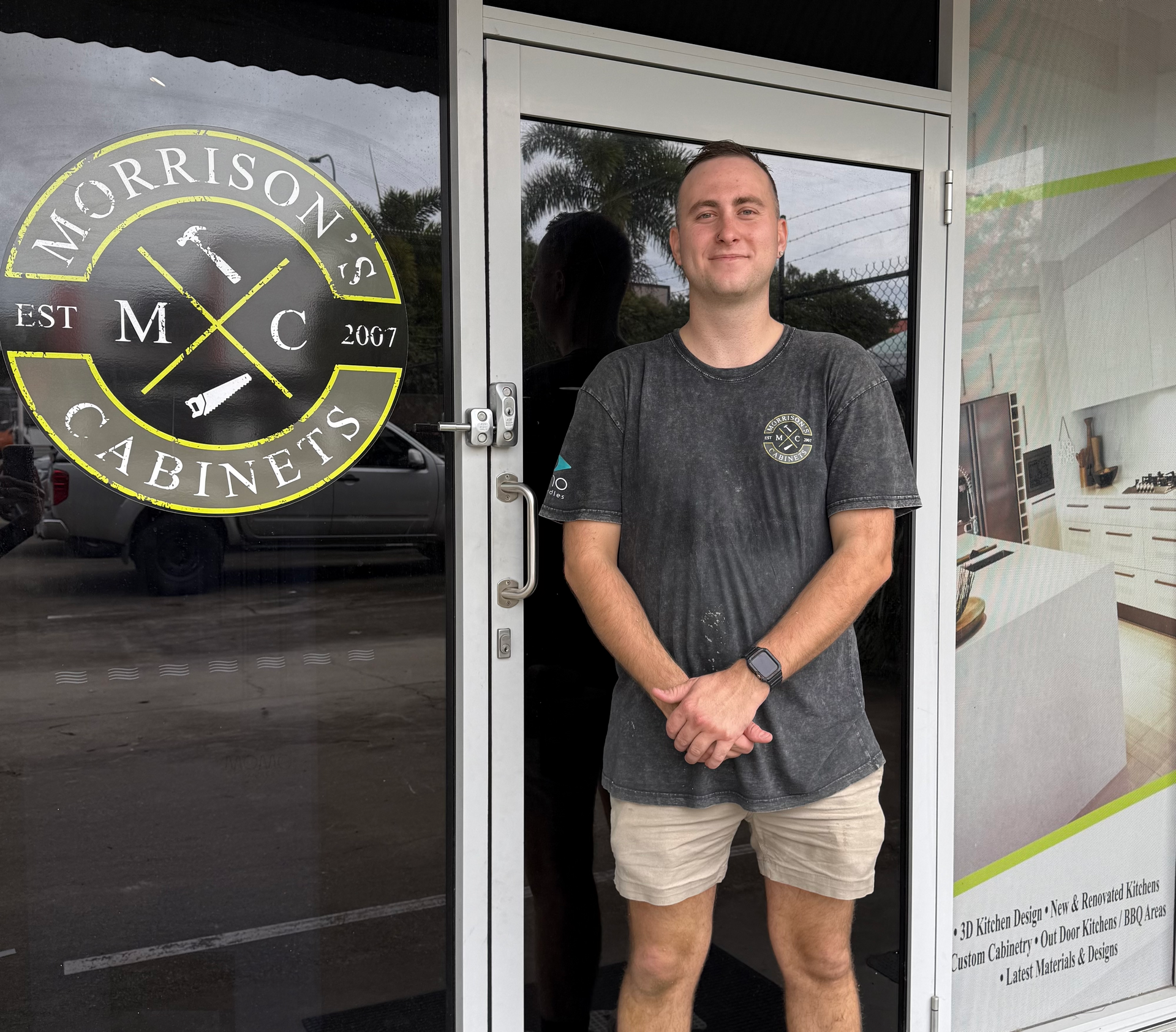 Man standing at a glass storefront with a Morrisons Cabinets logo, wearing a dark T-shirt and tan shorts.