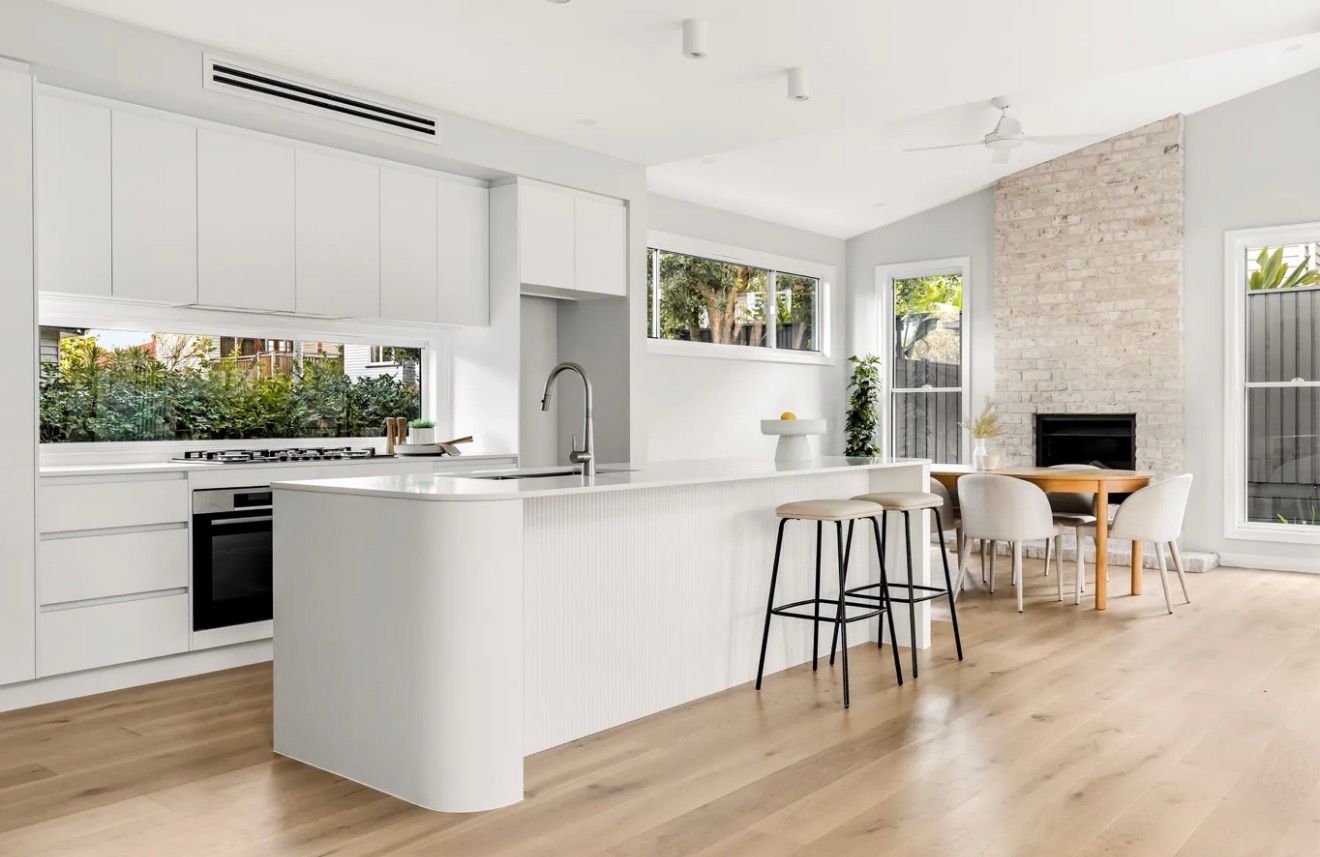 Bright white open-plan kitchen and dining area with wood floors and a small table by a fireplace