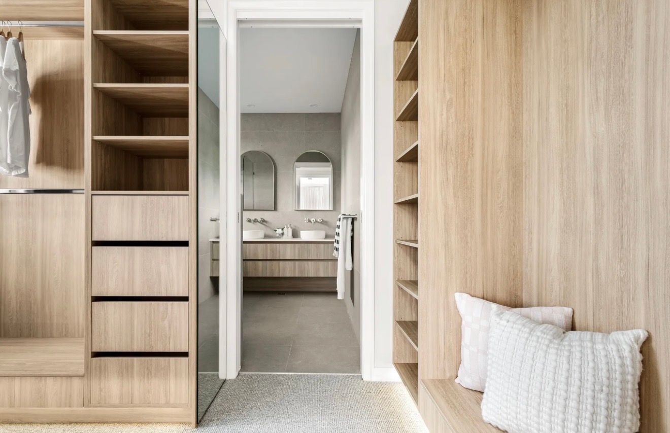 Modern walk-in closet with wood shelving, drawers, and a doorway to a bathroom; bench with pillows on the right