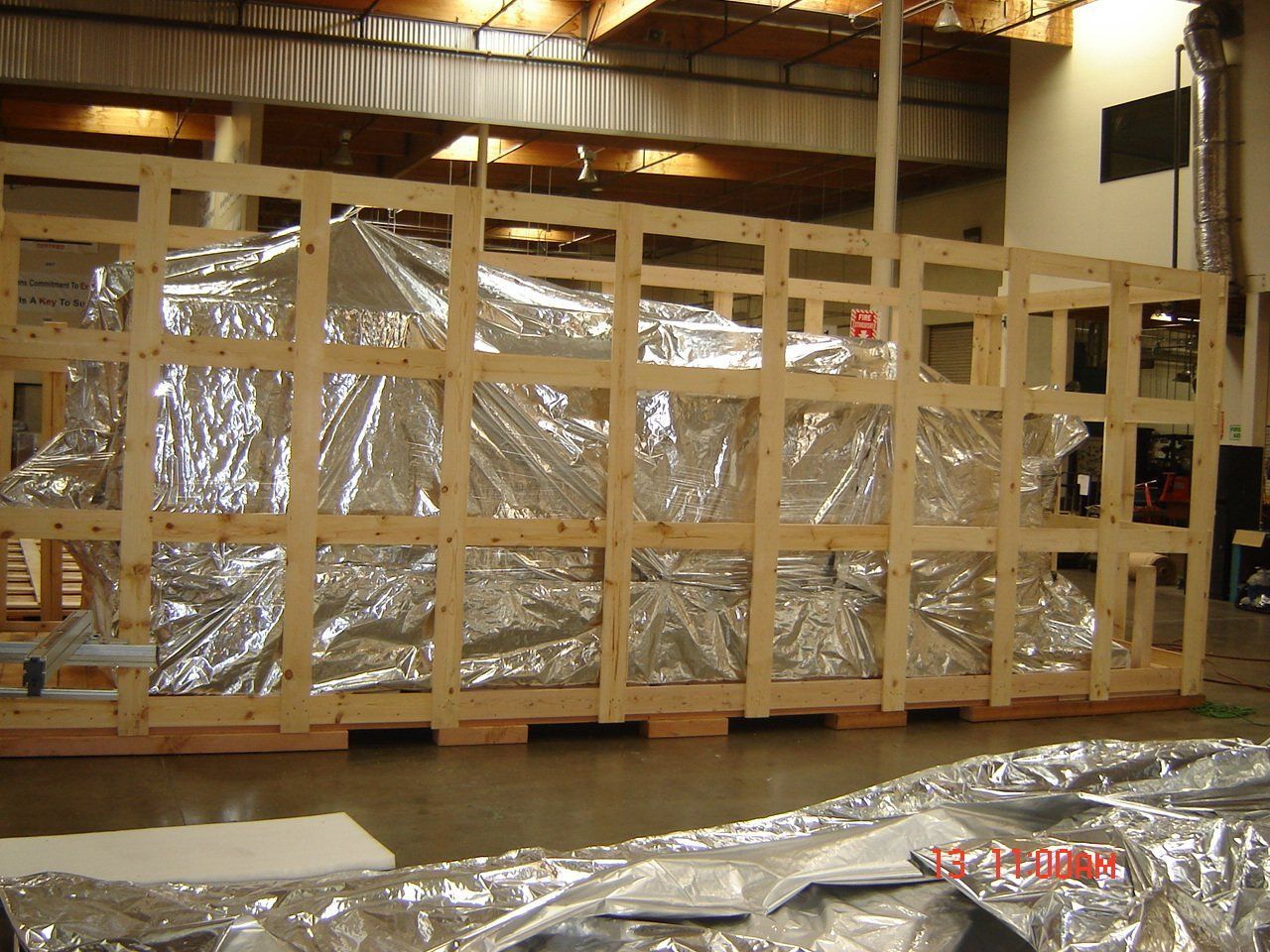 Large, wood-framed crate containing an object wrapped in reflective, silver material in an industrial setting.