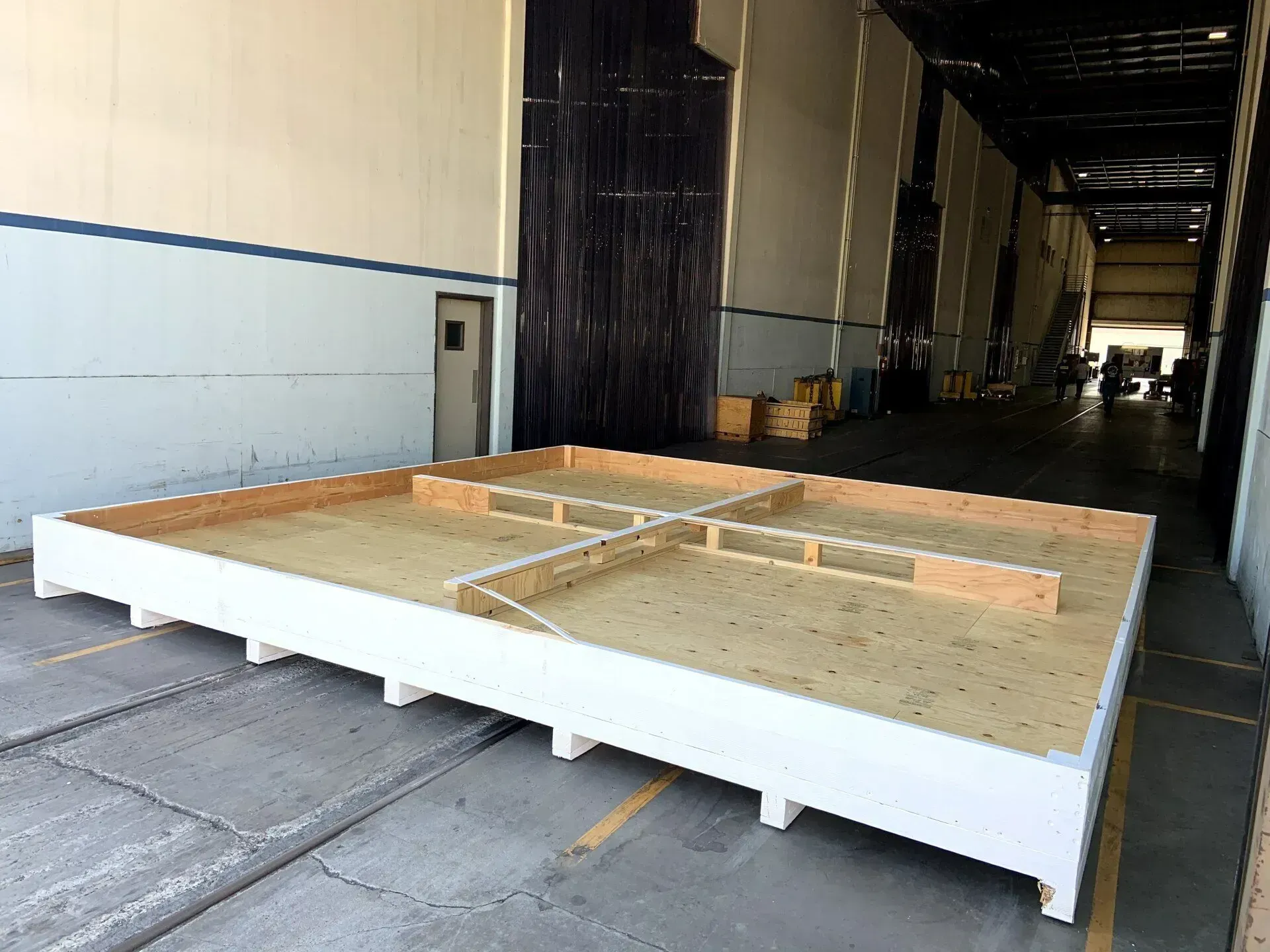A large, rectangular white wooden platform with interior dividers sits on a concrete floor inside a large warehouse.