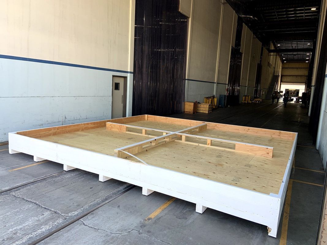 Large wooden platform with white frame and cross-shaped interior supports, inside a large warehouse.