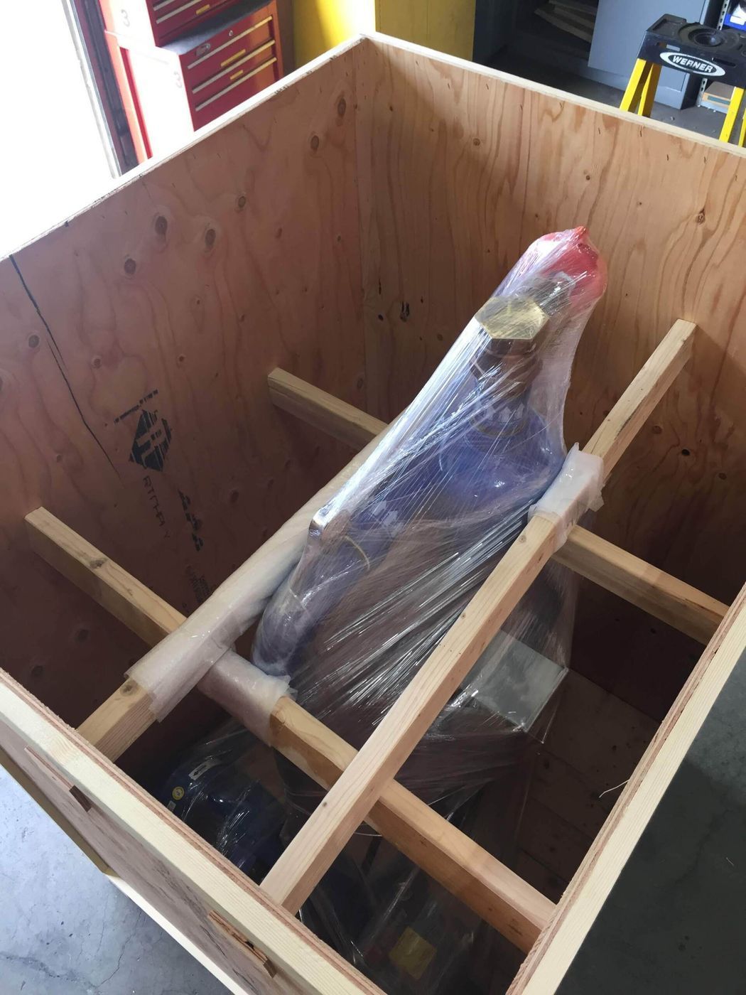 Blue object wrapped in plastic, secured inside a wooden crate with supporting beams.