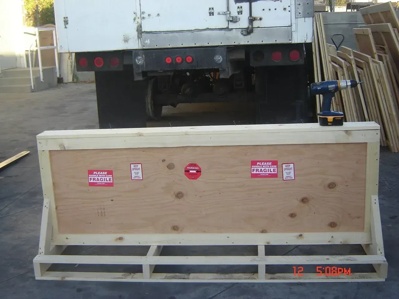 Wooden crate with shipping labels in front of a truck, a power drill is next to the crate.