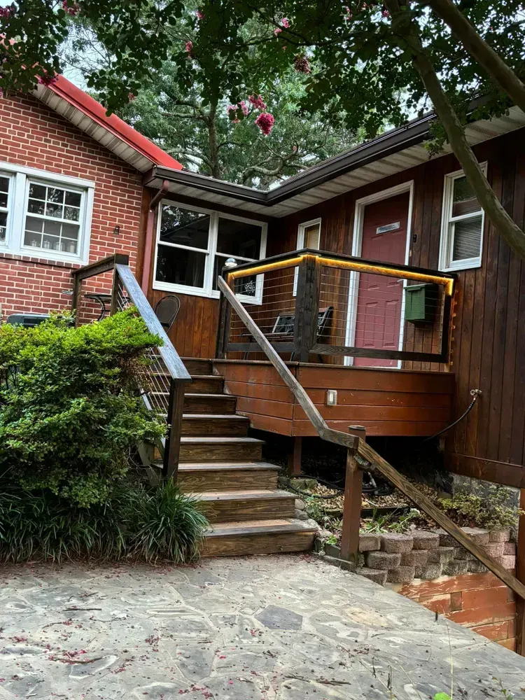 Brick house entrance with stairs, metal handrails, and a ramp beside a wooden porch under trees