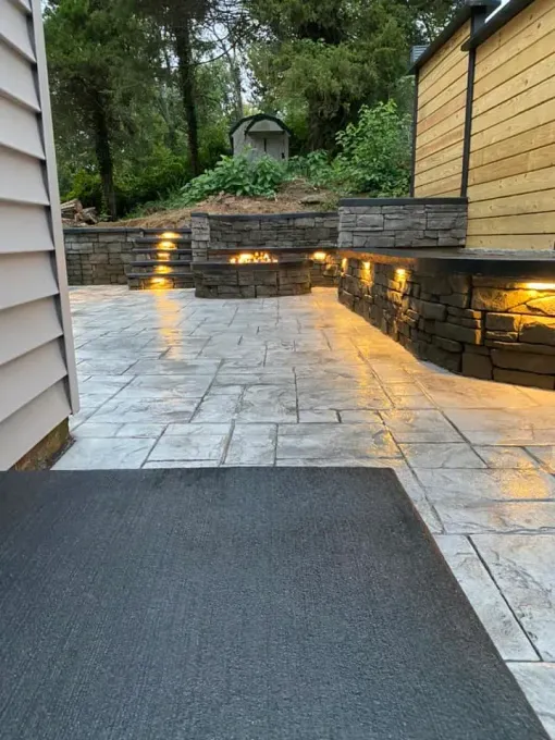 Stone patio with built-in steps and warm accent lights beside a house and garden trees.
