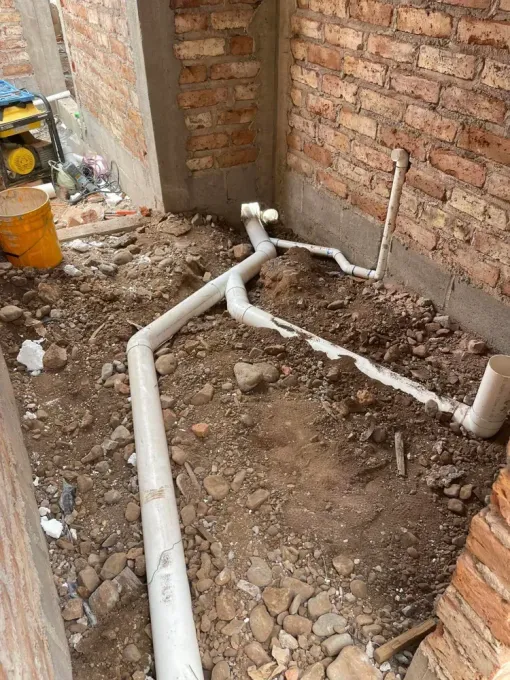 PVC plumbing pipes laid in a dirt trench beside a brick wall at a construction site