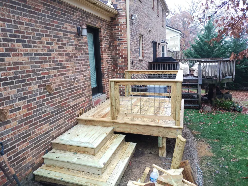 Wooden deck construction beside a brick house, with stacked lumber and a raised railing section.