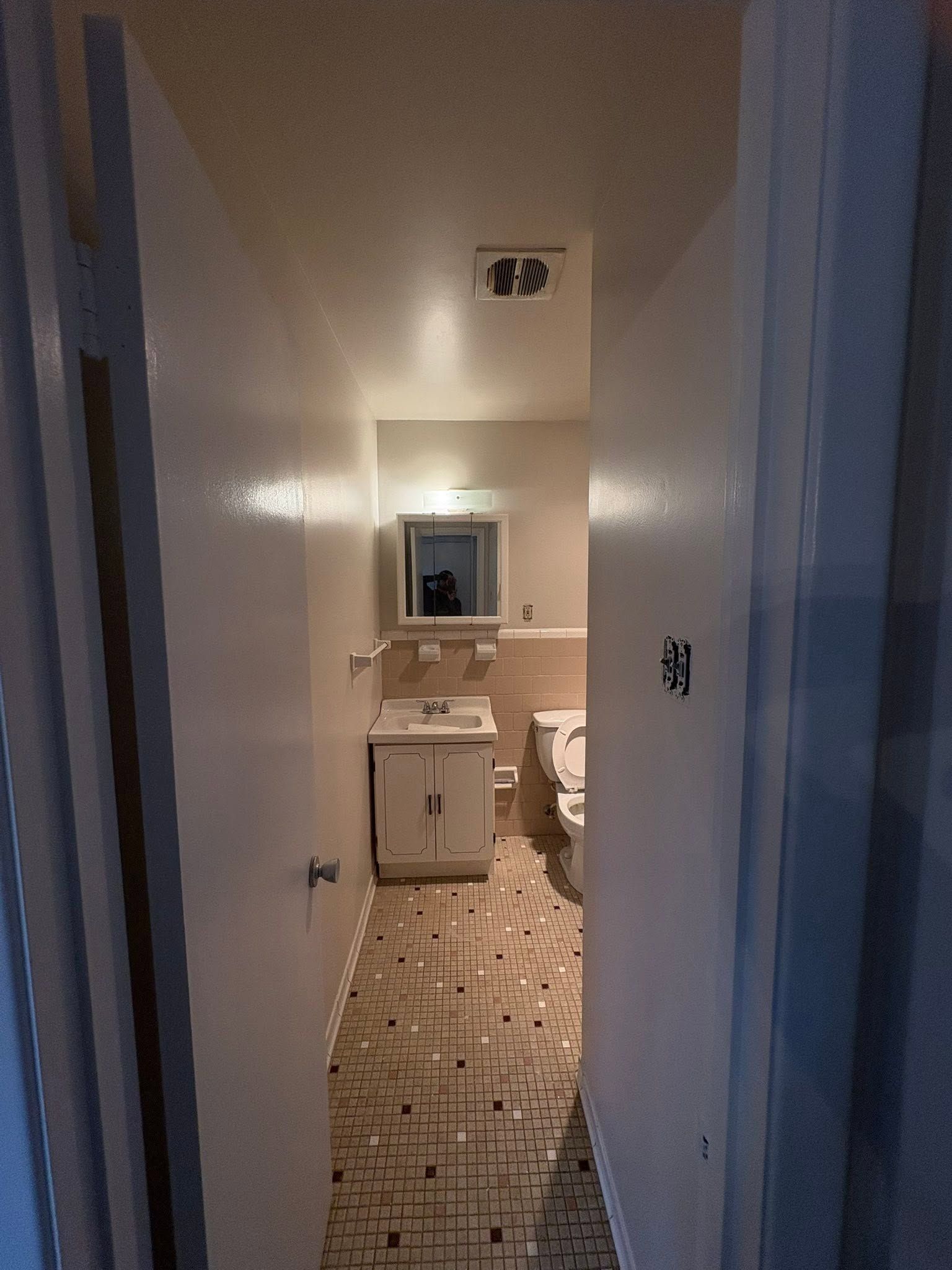 Narrow bathroom hallway with sink, mirror, toilet, and patterned tile floor.