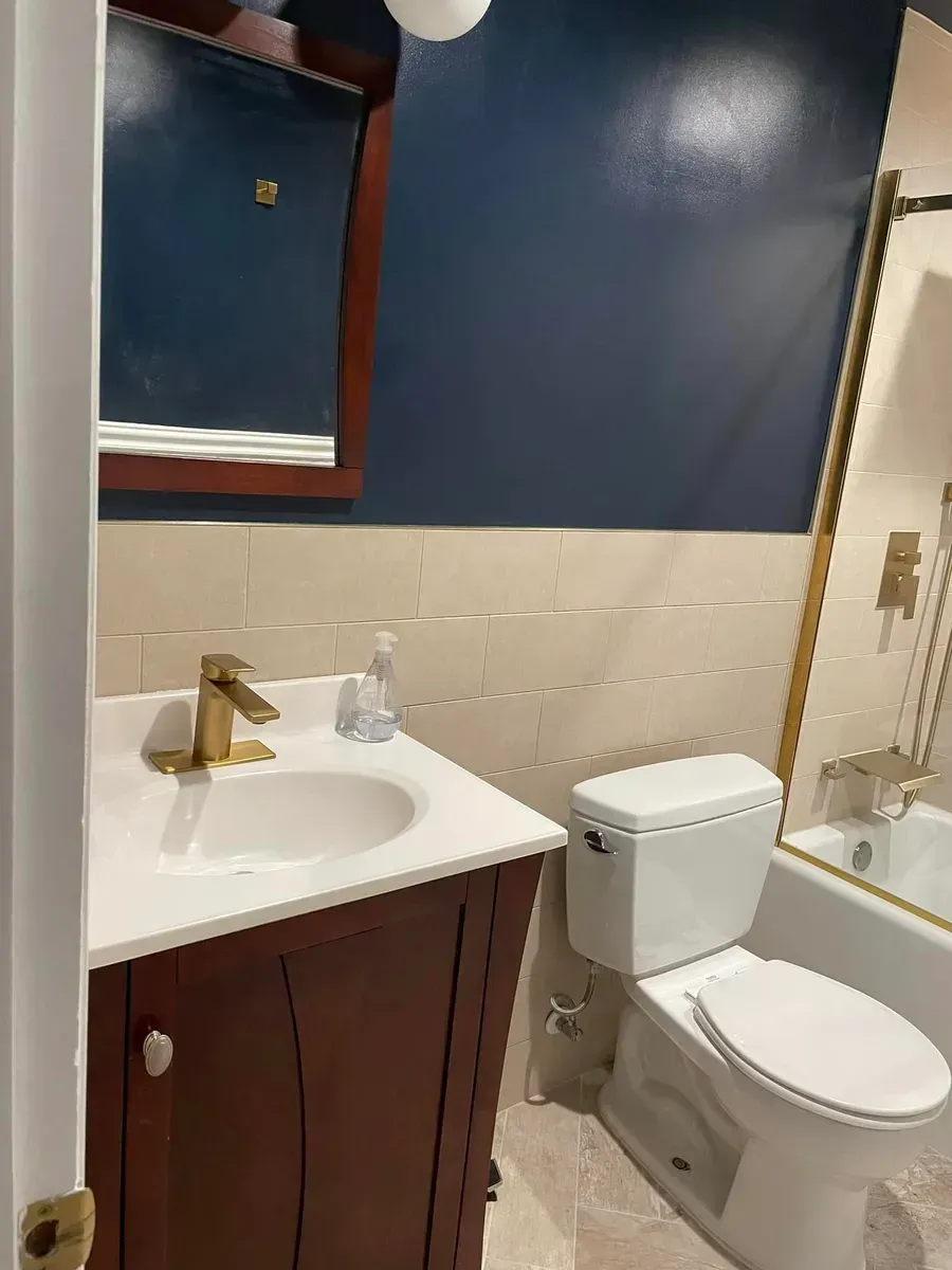 Small bathroom with white sink, gold faucet, toilet, blue walls, and tub shower on the right.