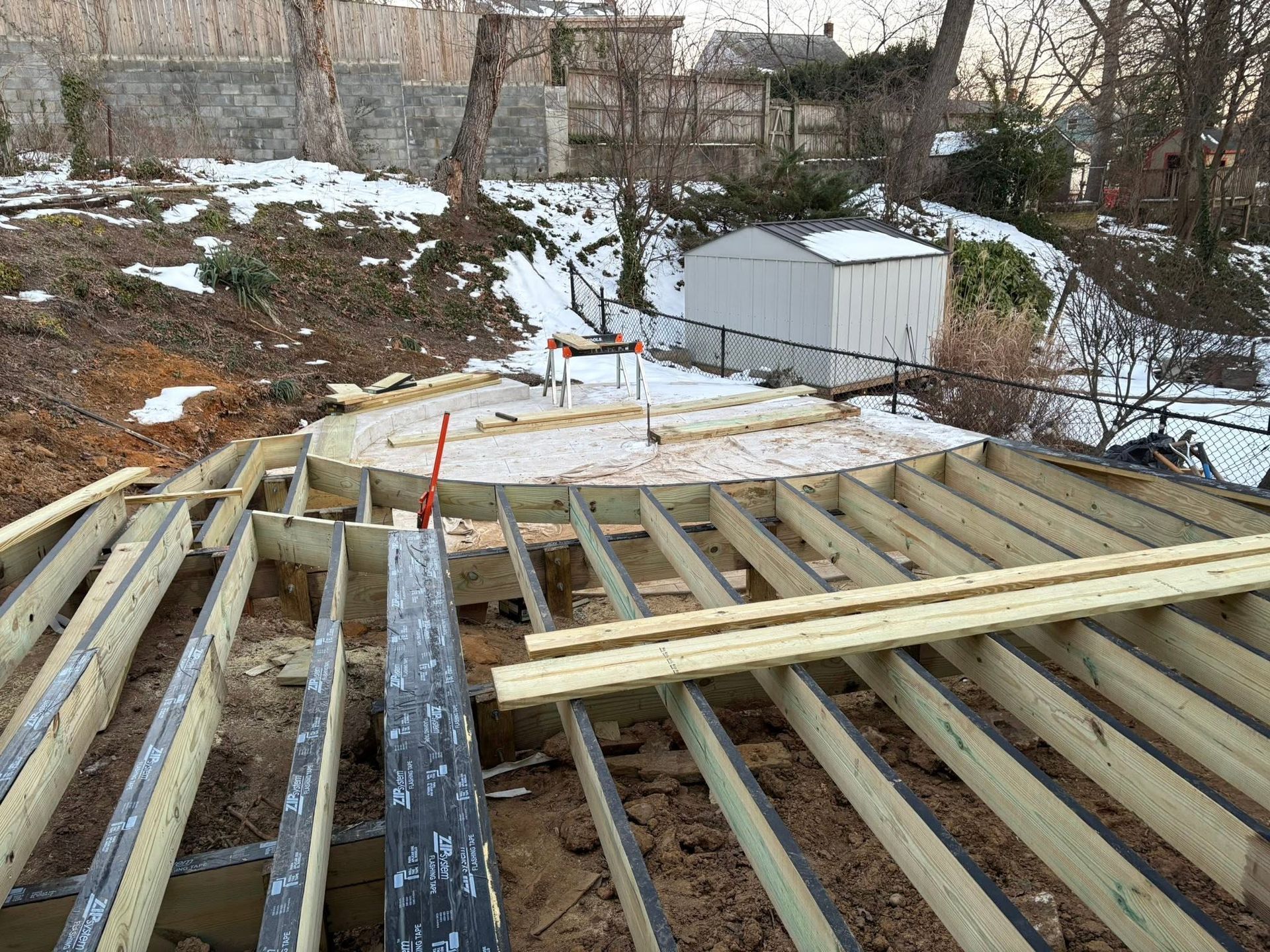 Partially built wooden roof deck overlooking a snowy backyard and sheds