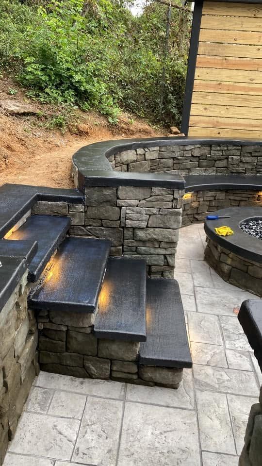 Stone patio with curved built-in seating, black steps, and a wooded hillside beside a house