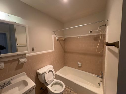 Small beige bathroom with sink, toilet, bathtub, mirror, and wall-mounted shower curtain rod.