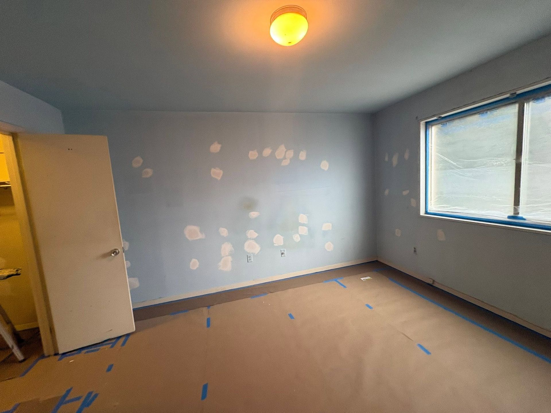 Empty room with light blue walls, beige carpet, a window, and a ceiling light; paint spots on one wall.