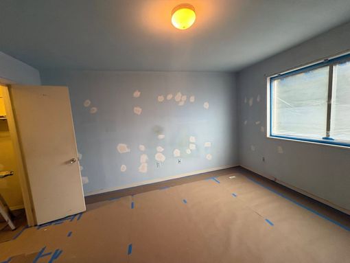 Empty room with light blue walls, beige carpet, a window, and a ceiling light; paint spots on one wall.